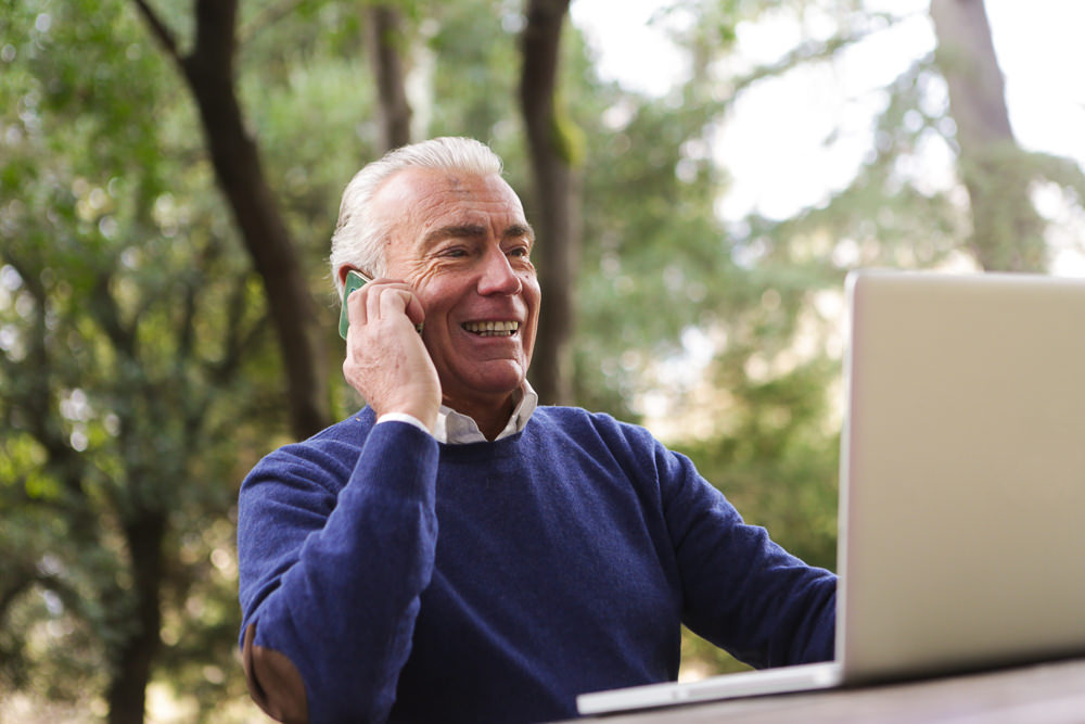 Senior man has a personal conversation on his cell phone while sitting in front of his open laptop