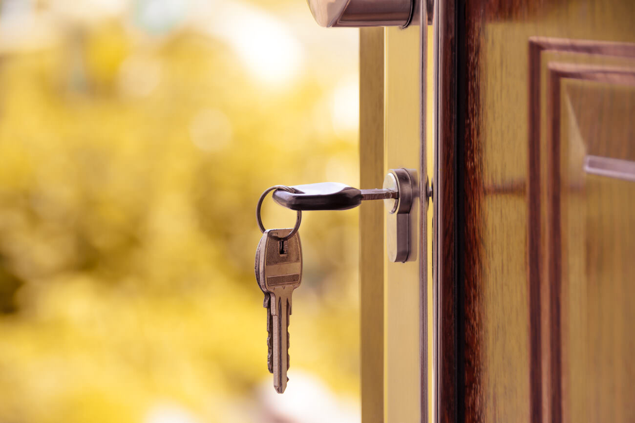 A key hangs from a lock on a door. The door is open, looking outwards, and it is bright and sunny outside with trees in the background. Someone is moving out of their house in November