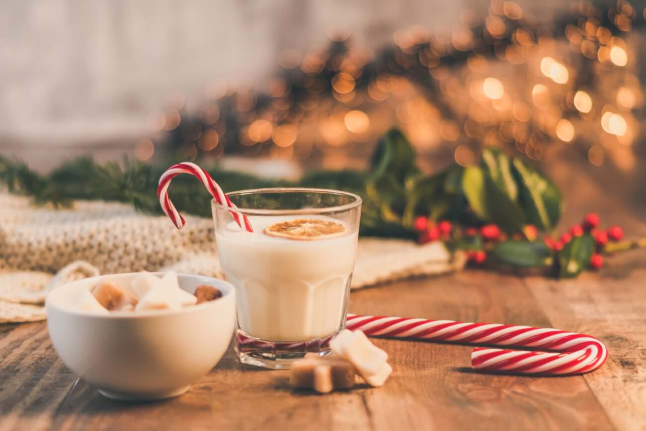 Cookies and a glass of milk sit next to a candy cane on a table. Warm lighting in the background reveals dark green holly leaves and red berries